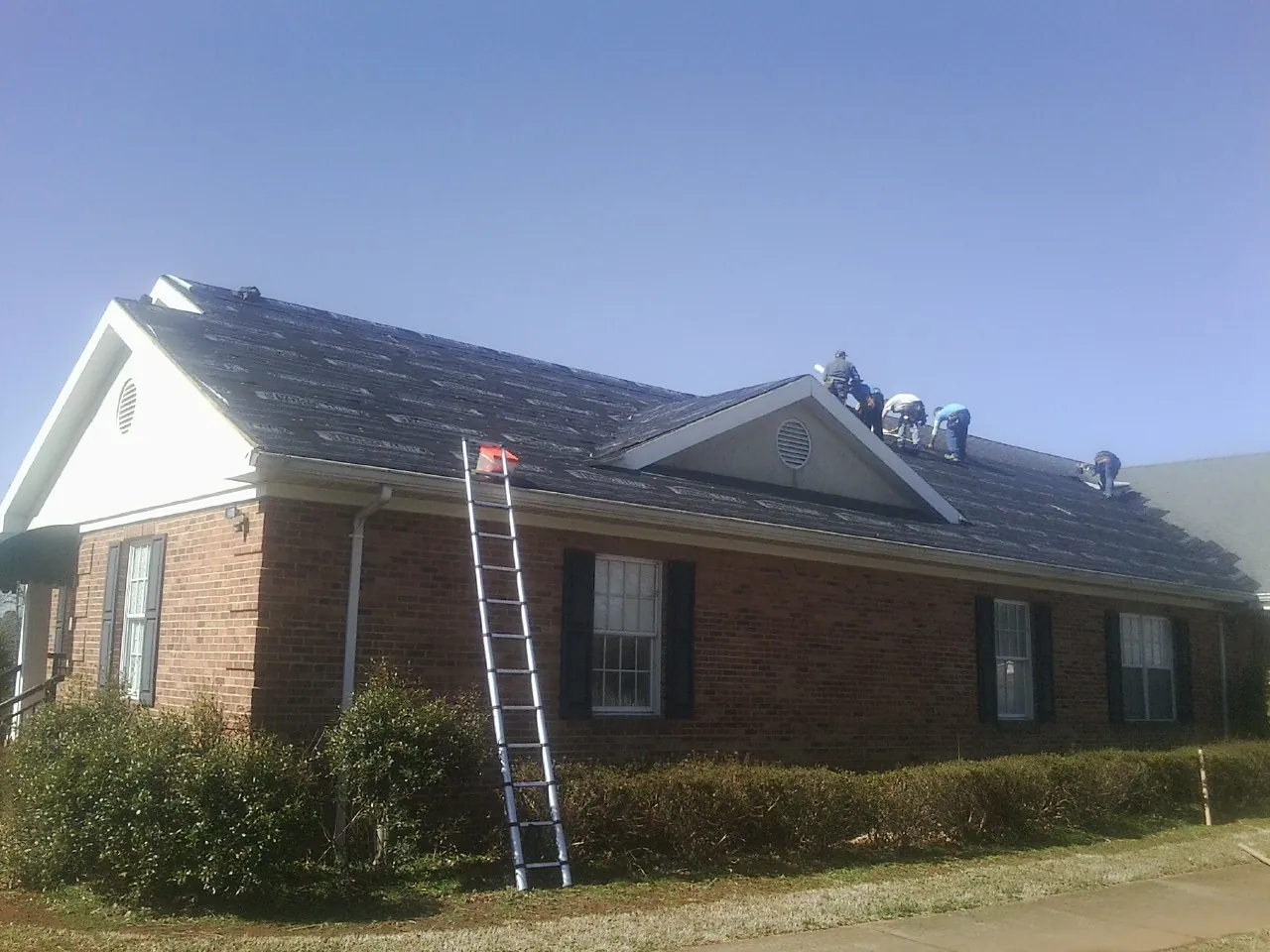 Crew tearing shingles off a brick ranch roof with a ladder set — Emergency Roof Repair work by our crew in New Waverly